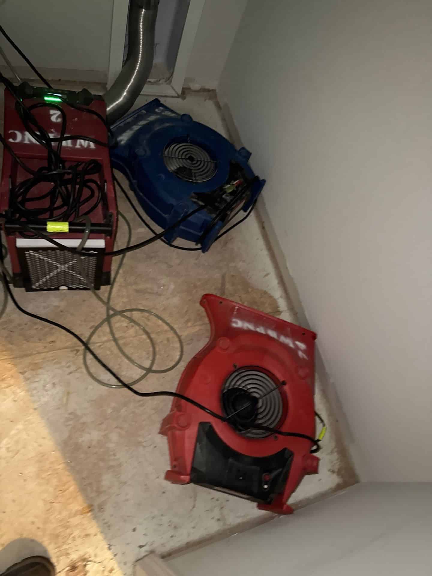 Water Damage Restoration Equipment Including Air Movers And A Dehumidifier Set Up In A Residential Corner To Dry Subfloor And Prevent Further Moisture-Related Damage.